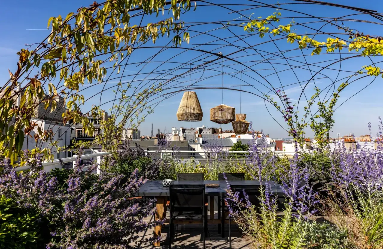 Une pergola au milieu du jardin, avec une table et des chaises, des fleurs violettes et des lanternes suspendues, offre une vue sur les toits de Paris.