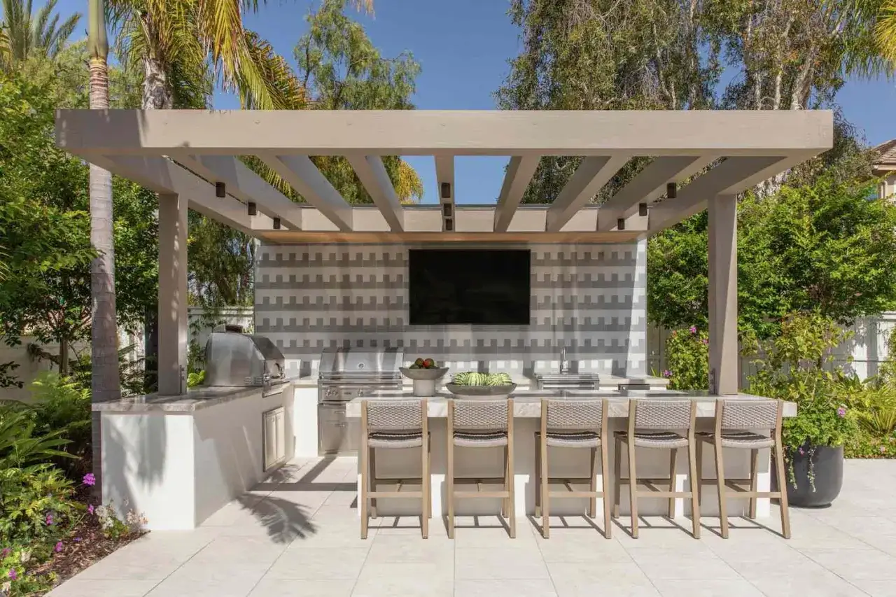 Une cuisine ext&eacute;rieure moderne sous une pergola non couverte, avec un grand barbecue, un bar et des tabourets.