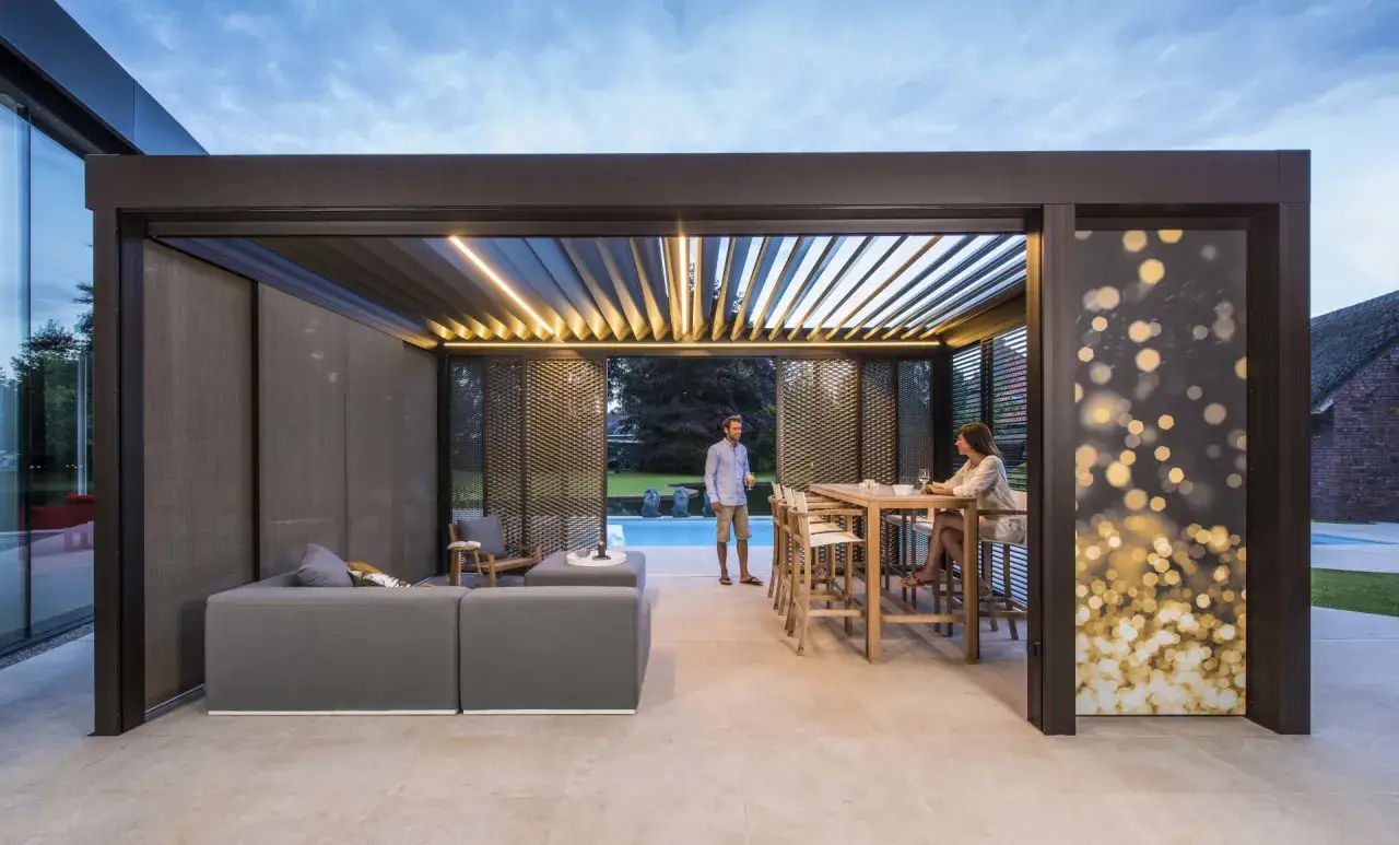 Un couple profite d'une soirée sous une pergola bioclimatique. La hauteur de la structure offre un espace de vie extérieur confortable avec une piscine en arrière-plan.