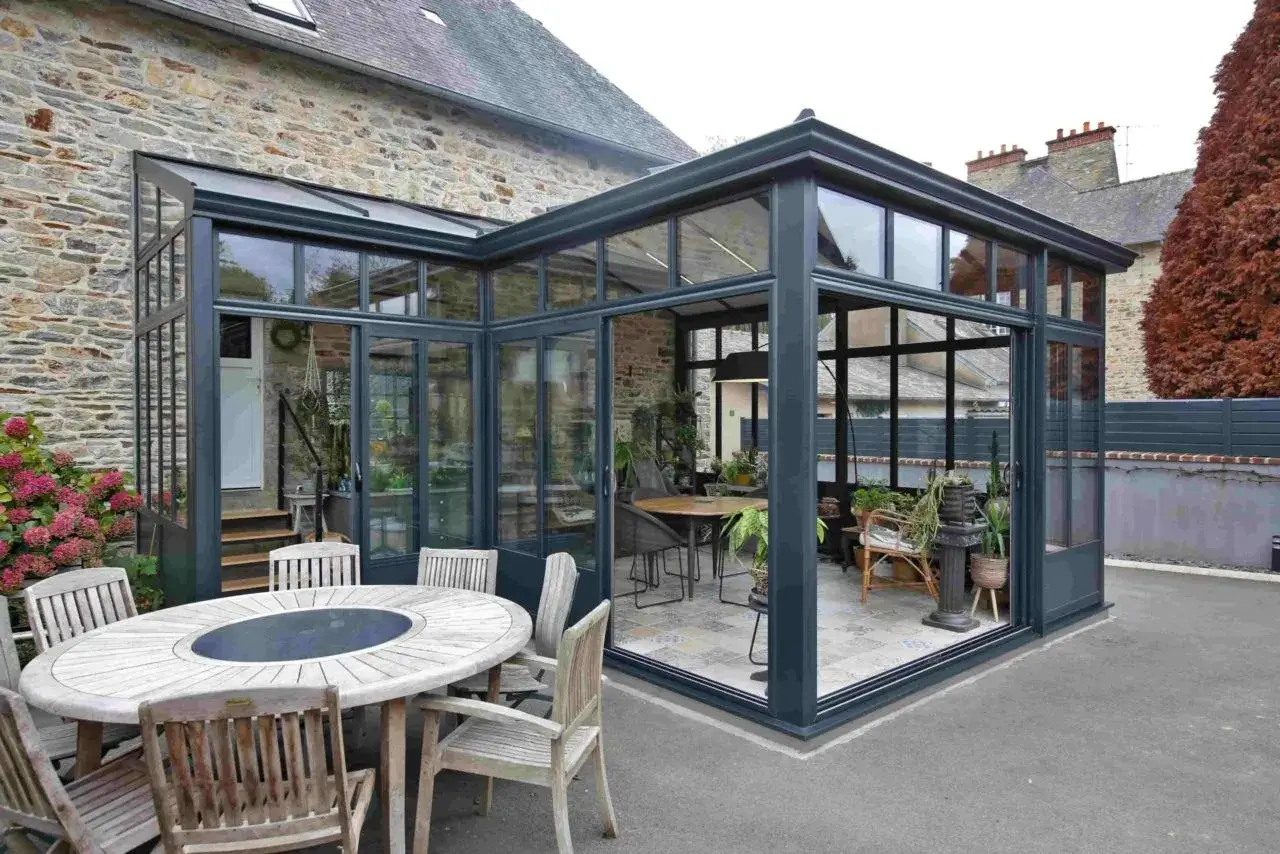 Plan de v&eacute;randa &agrave; faire soi-m&ecirc;me : une structure moderne en verre et m&eacute;tal s'int&egrave;gre &agrave; une maison en pierre, avec une table ext&eacute;rieure et des plantes.
