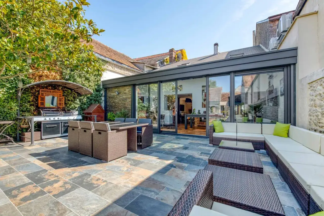 Terrasse en pierre avec salon de jardin, barbecue et une veranda sans permis de construire aux grandes baies vitrées.
