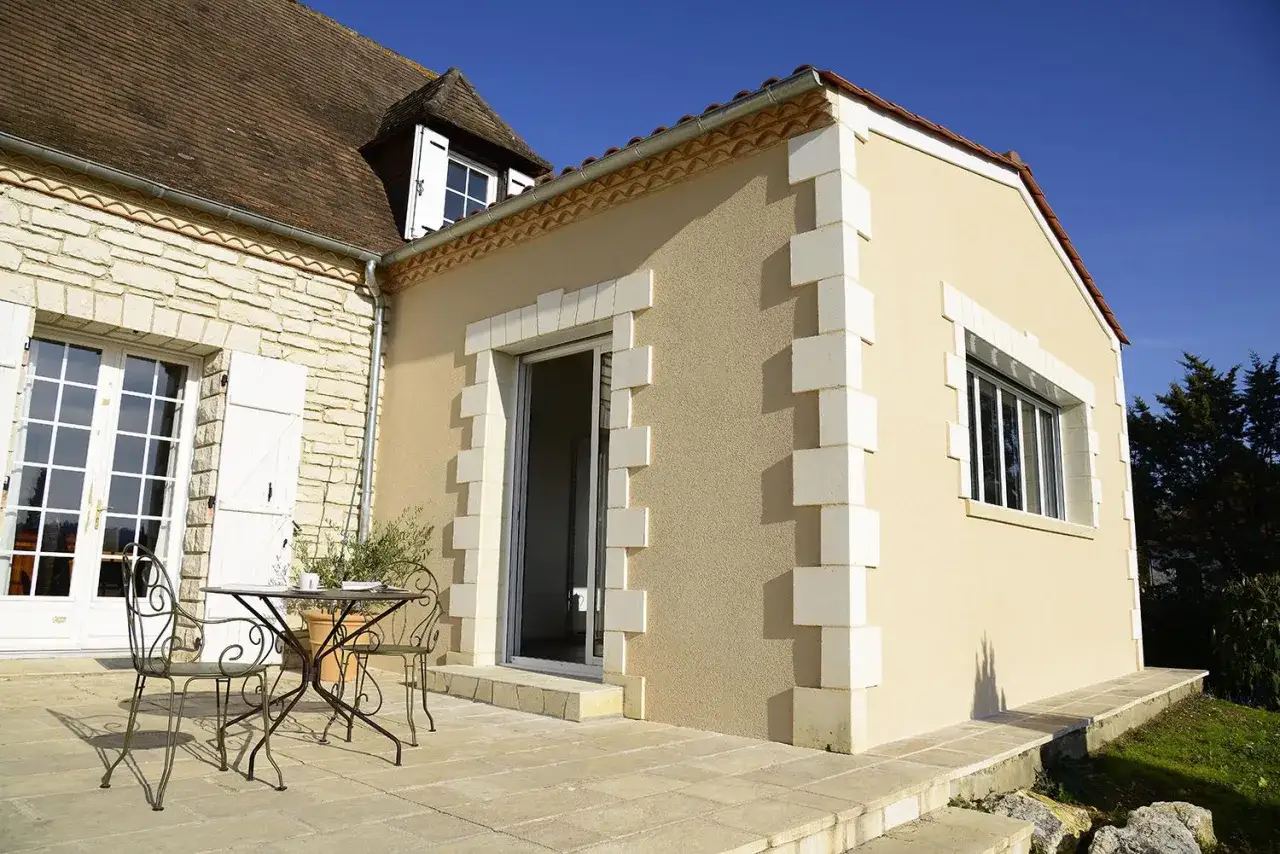 Extension de maison sur terrasse b&eacute;ton. Une table et des chaises invitent &agrave; profiter du soleil.