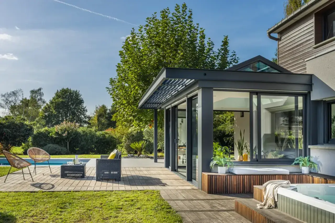 Une pergola bioclimatique moderne prolonge une maison, offrant un espace de vie extérieur avec une piscine et un jacuzzi.