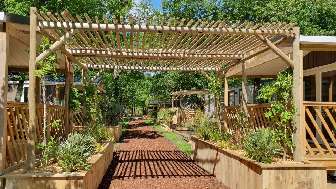 All&eacute;e bord&eacute;e de jardini&egrave;res en bois sous un toit pergola naturel, menant &agrave; des h&eacute;bergements de vacances.