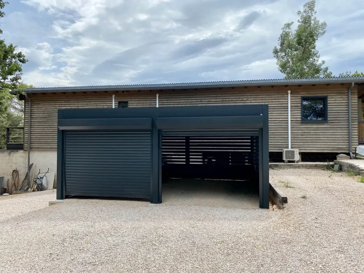 Un carport TVA moderne avec un volet roulant ferm&eacute; et un espace ouvert, adoss&eacute; &agrave; une maison en bois.