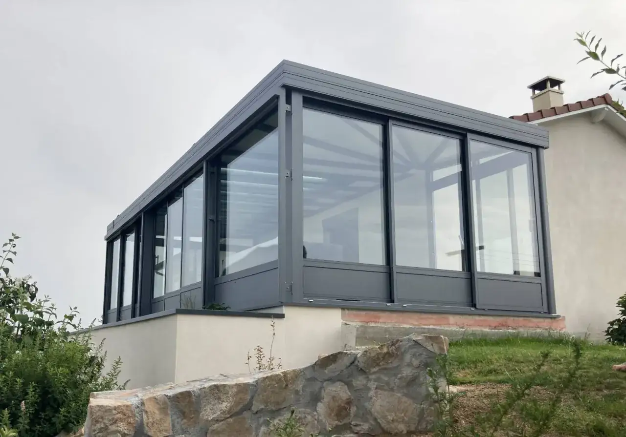 Moderne veranda adoss&eacute;e au mur, avec de grandes baies vitr&eacute;es coulissantes en aluminium gris. Un espace lumineux pour profiter du jardin.