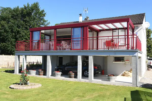 Une maison avec une veranda terrasse rouge vif, offrant une vue sur un jardin verdoyant et une voiture garée sous le balcon.