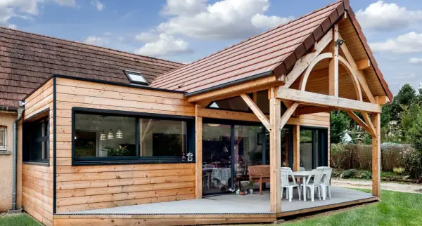 Extension de maison en bois avec terrasse sur dalle béton. Une table et des chaises sont installées sous un auvent.