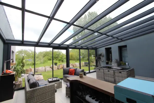 Salon moderne avec un toit en verre laissant entrer la lumière. Des canapés confortables invitent à la détente, tandis que le jardin verdoyant offre une vue paisible.
