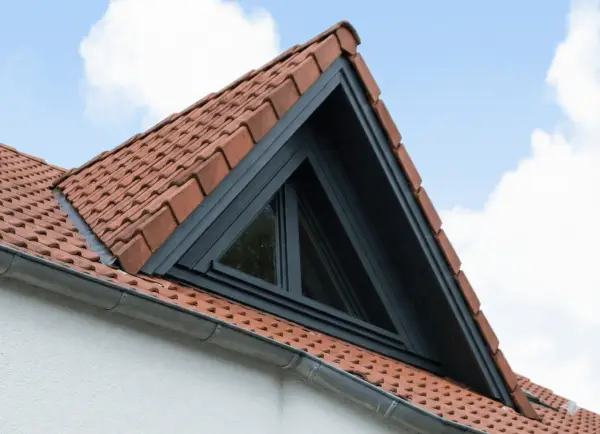 Un puit de lumière triangulaire s'intègre dans un toit en tuiles rouges, sous un ciel bleu et nuageux.