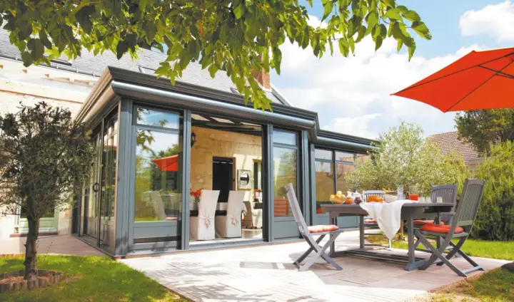 Une véranda moderne avec un surbot béton, une table et des chaises sur une terrasse en bois, sous un parasol orange vif.