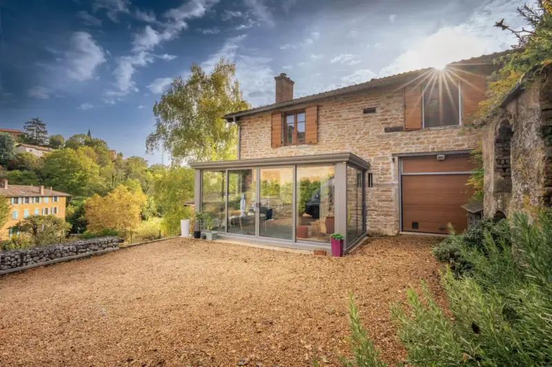 Maison en pierre avec une veranda lumineuse, un jardin de gravier et un paysage vallonné en automne.
