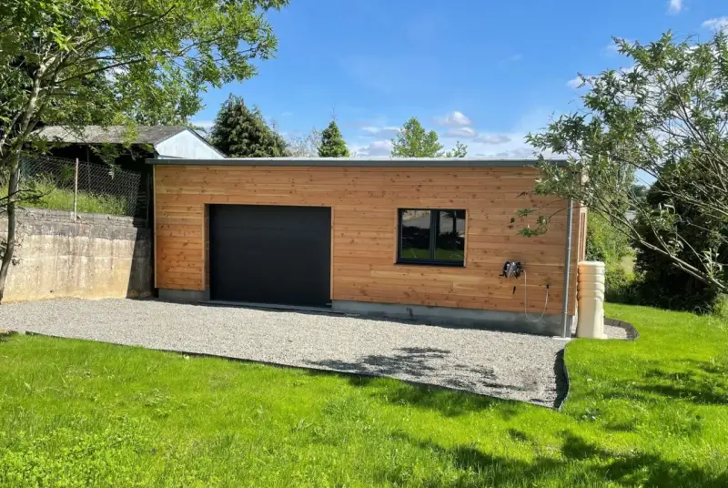 Extension garage bois accolé maison, avec porte noire, fenêtre et jardin verdoyant.